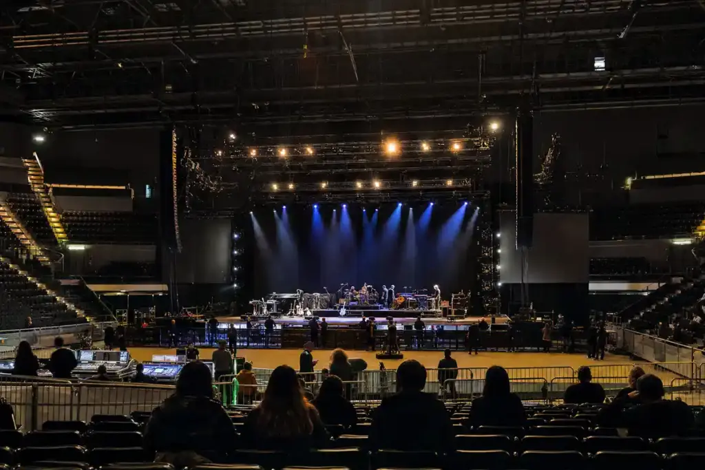 Palasport con palco in allestimento, luci blu accese e persone in silhouette tra platea e transenne