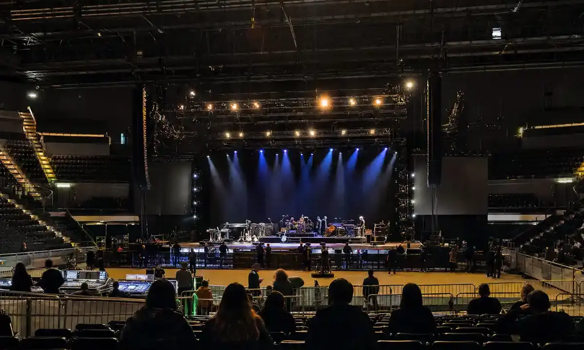 Palasport con palco in allestimento, luci blu accese e persone in silhouette tra platea e transenne