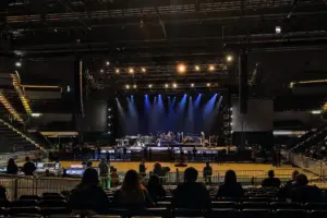 Palasport con palco in allestimento, luci blu accese e persone in silhouette tra platea e transenne