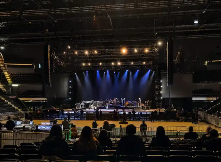 Palasport con palco in allestimento, luci blu accese e persone in silhouette tra platea e transenne