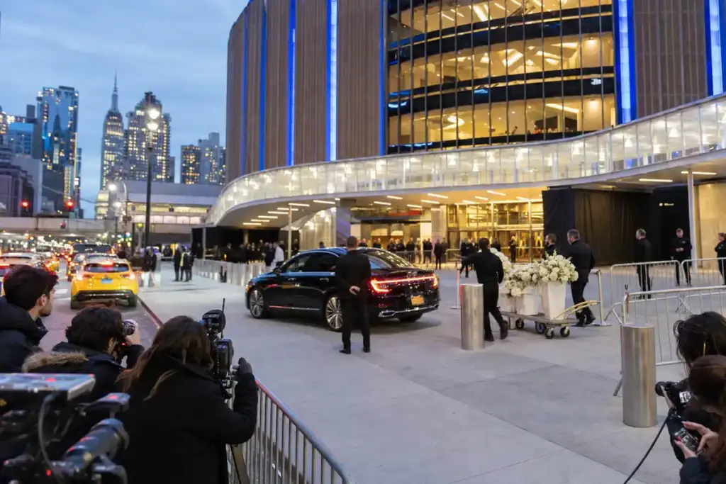 Ingresso di una grande arena a New York al tramonto, con fotografi, sicurezza, auto di lusso e carrello di fiori bianchi