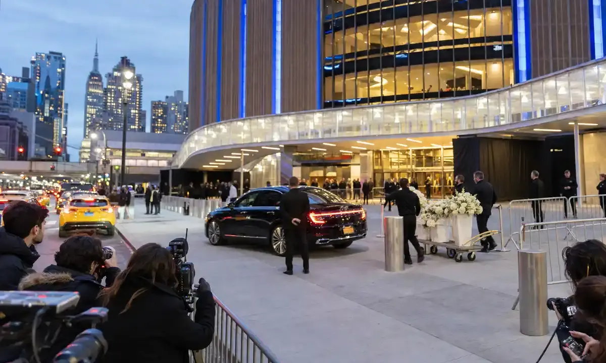 Ingresso di una grande arena a New York al tramonto, con fotografi, sicurezza, auto di lusso e carrello di fiori bianchi