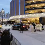 Ingresso di una grande arena a New York al tramonto, con fotografi, sicurezza, auto di lusso e carrello di fiori bianchi