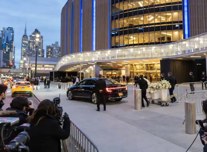 Ingresso di una grande arena a New York al tramonto, con fotografi, sicurezza, auto di lusso e carrello di fiori bianchi