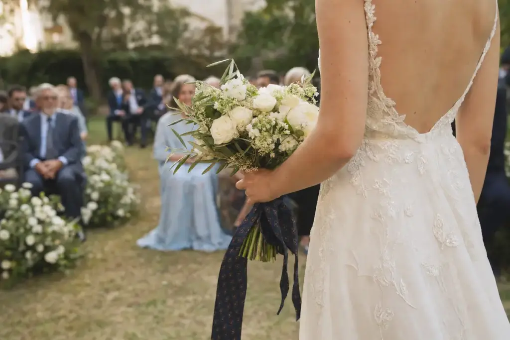 Sposa di spalle con abito ricamato e bouquet di fiori bianchi legato con una cravatta scura, invitati sfocati in giardino