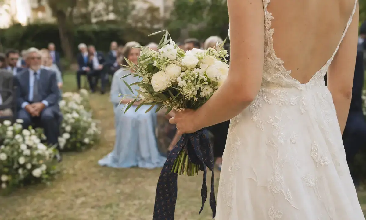 Sposa di spalle con abito ricamato e bouquet di fiori bianchi legato con una cravatta scura, invitati sfocati in giardino