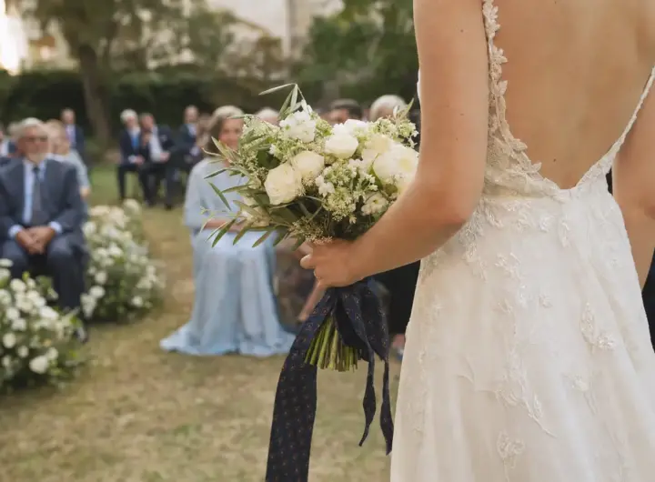 Sposa di spalle con abito ricamato e bouquet di fiori bianchi legato con una cravatta scura, invitati sfocati in giardino