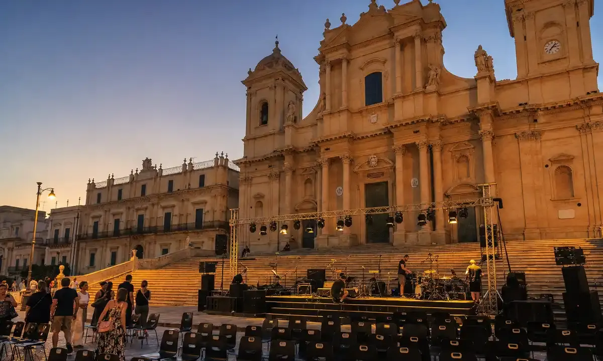Palco in allestimento davanti a una cattedrale barocca al tramonto, con tecnici e file di sedie vuote in piazza