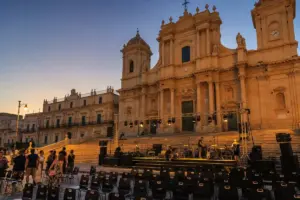 Palco in allestimento davanti a una cattedrale barocca al tramonto, con tecnici e file di sedie vuote in piazza