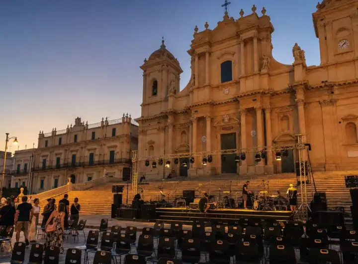 Palco in allestimento davanti a una cattedrale barocca al tramonto, con tecnici e file di sedie vuote in piazza